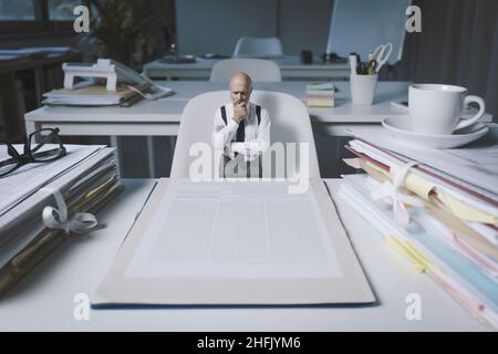 Piccolo uomo d'affari che controlla un contratto enorme di affari, sta pensando con la mano sul mento Foto Stock