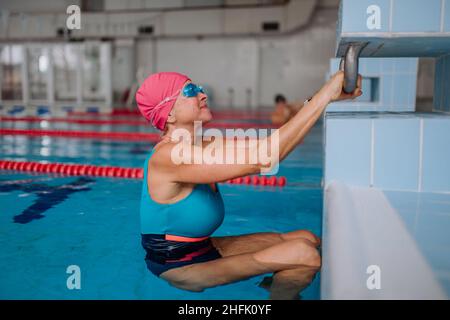 Nuotatore donna anziano attivo che tiene sul blocco di partenza preparandosi a nuotare nella piscina interna. Foto Stock