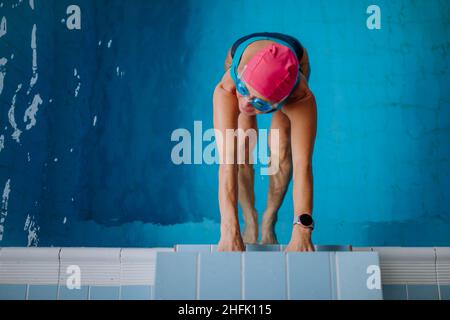 Vista dall'alto di una donna attiva che tiene il nuotatore sul blocco di partenza nella piscina interna. Foto Stock