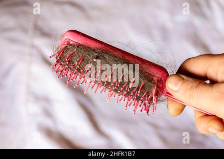 Capelli grigi e scuri su un pettine rosso in mani maschili come conseguenza di malattia o mancanza di vitamine e minerali nel corpo, fuoco selettivo Foto Stock