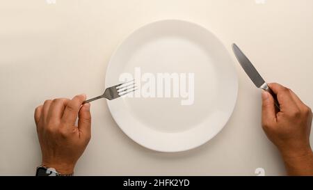 Immagine vista dall'alto di un uomo che tiene la forcella e il coltello sopra il tavolo da pranzo. Piastra bianca in ceramica. Svuotare il mockup della piastra Foto Stock