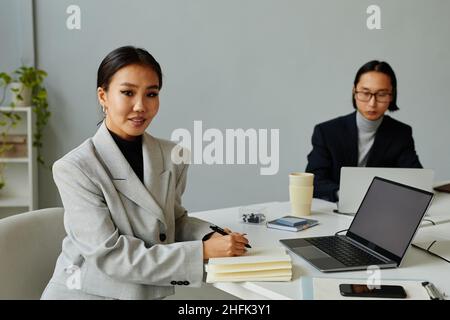 Ritratto della giovane donna d'affari asiatica che guarda la macchina fotografica e sorride mentre lavora al tavolo in un ufficio minimal moderno Foto Stock