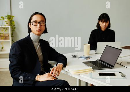 Ritratto del giovane uomo d'affari asiatico guardando la fotocamera con serio volto espressione mentre si lavora a tavola in un ufficio moderno minimo, spazio copia Foto Stock