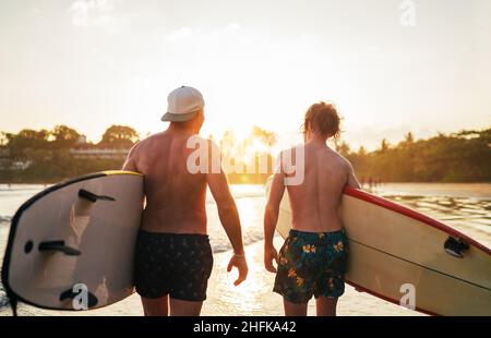 Padre con figlio adolescente che cammina con tavole da surf presso la spiaggia sabbiosa dell'oceano con palme sullo sfondo illuminate dal sole al tramonto. Sorridevano ed ha Foto Stock