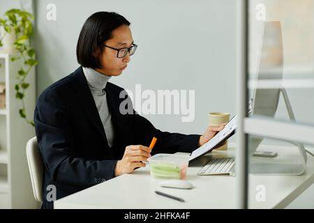 Ritratto della vista laterale dell'uomo d'affari asiatico che mangia spuntini sani mentre legge i documenti in ufficio Foto Stock