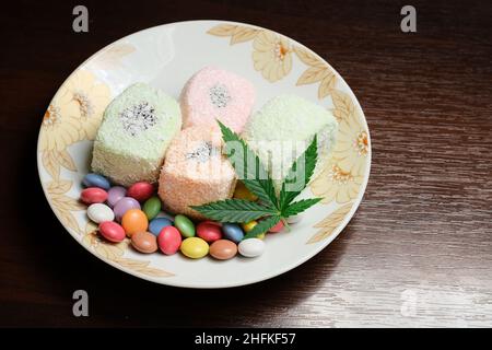 Dessert di cannabis. Delizia turca con fiocchi di cocco e confetti di caramelle colorate, vista dall'alto. Foglie verdi di cmarijuana su sfondo bianco della piastra. Layout Foto Stock