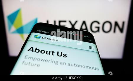 Telefono cellulare con sito web della società di software svedese Hexagon AB sullo schermo di fronte al logo aziendale. Mettere a fuoco sulla parte superiore sinistra del display del telefono. Foto Stock