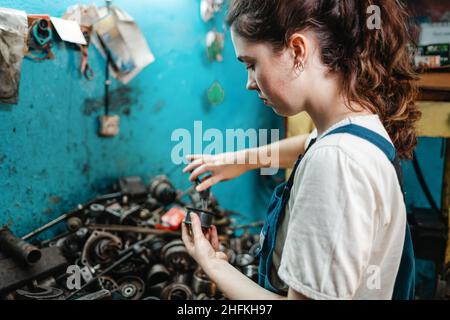 Parità di genere. Ritratto di una giovane donna bruna in uniforme, impegnata nella riparazione di parti di auto. Parete blu e parti di ricambio sullo sfondo. Foto Stock