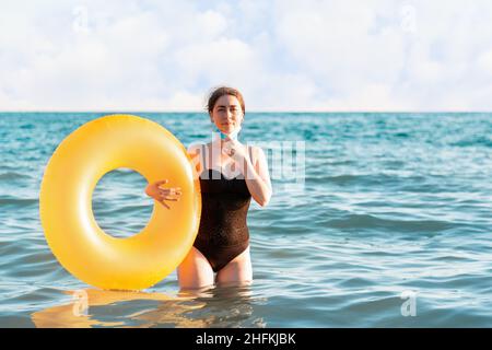 Distanza sociale e protezione dai virus. Una donna in costume da bagno rimuove una maschera medica e si pone con un cerchio gonfiabile mentre si trova in mare. Foto Stock