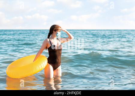 Distanza sociale e protezione dai virus. Una donna in costume da bagno e maschera medica si pone con un cerchio gonfiabile in piedi nel mare e guarda via. IO Foto Stock