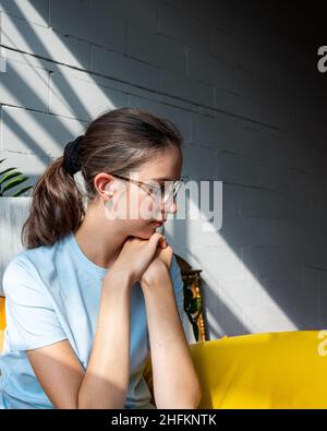 Ragazza adolescente sconvolto in occhiali e abiti casual che soffrono di solitudine guarda giù su uno sfondo con ombre diagonali, vista laterale, gram verticale Foto Stock