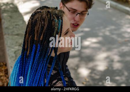 Una giovane coppia eterosessuale chiacchierando mentre si siede su una panchina in un parco cittadino. Una donna con lunghi lucchetti blu. L'uomo che indossa gli occhiali. Selettivo Foto Stock