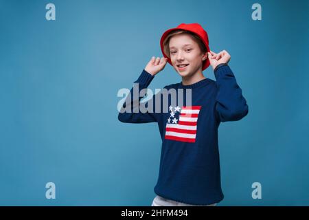 Ritratto di un attraente giovane maschio caucasico. Bel ragazzo adolescente in maglione blu con noi bandiera e cappello rosso. Ragazzo sorridente che si posa in studio su sfondo blu. Foto Stock