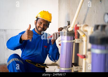 smillando il giovane ingegnere indiano o riparatore che mostra i pollici in su segno o gesto della mano mentre lavora al luogo di costuazione - concetto di blu professionale Foto Stock