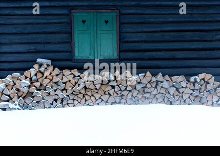 Una finestra con persiane verdi con cuori su una vecchia cabina alpina in inverno con neve e legna da ardere Foto Stock