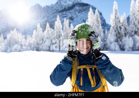 Ritratto di ragazzo sorridente carino con le mani sulle guance, scarpetta parzialmente coperta di neve nel paesaggio invernale nella valle di Krnica, Slovenia Foto Stock