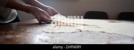 Vista ad angolo basso delle mani femminili che tirano pasta vegana fatta in casa per uno strudel tradizionale su un tavolo da pranzo. Foto Stock