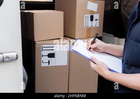 Vista ritagliata della scrittura del corriere sulla clipboard vicino a scatole di cartone sfocate in auto Foto Stock