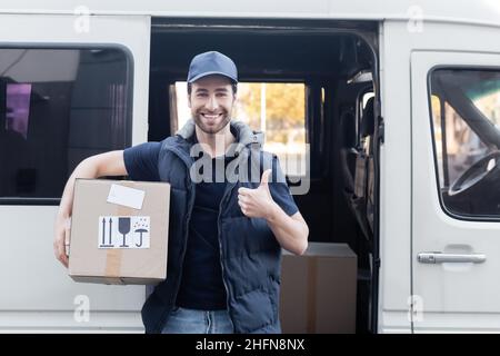 Corriere sorridente che mostra come e tenendo la scatola di cartone vicino all'auto all'aperto Foto Stock