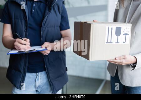 Vista ritagliata della scrittura del corriere sulla clipboard vicino alla fidanzata con scatola di cartone nel corridoio Foto Stock