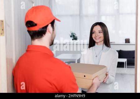 Donna positiva che prende le scatole della pizza dal corriere sfocato nel corridoio Foto Stock