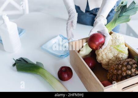 Vista ritagliata della donna in guanti di lattice che tengono la mela vicino al cibo fresco e antisettico a casa Foto Stock