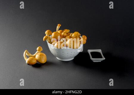 Deliziosi funghi con tappo dorato su sfondo nero in una ciotola bianca con una ciotola bianca di salsa di soia, fresca e cruda, da vicino Foto Stock