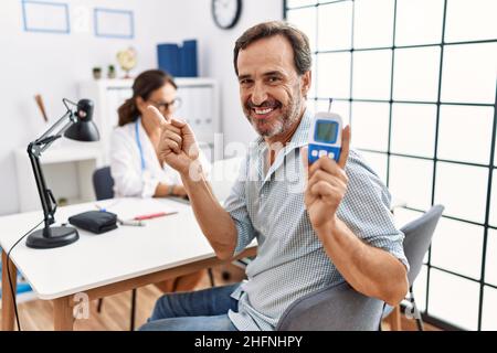 Uomo di mezza età al medico clinica tenendo dispositivo glucometro sorridente felice di puntamento con la mano e il dito al lato Foto Stock