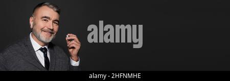 uomo anziano allegro che tiene gli auricolari mentre ascolta la musica isolato su nero, banner Foto Stock