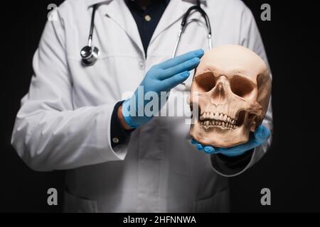 vista ritagliata del medico sfocato in guanti di lattice che tengono il cranio isolato su nero Foto Stock