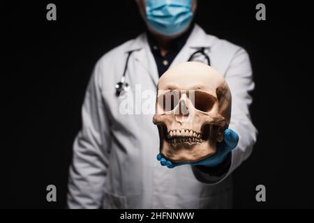vista ritagliata di medico sfocato in maschera medica tenendo cranio isolato su nero Foto Stock