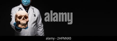 vista ritagliata di medico sfocato in maschera medica tenendo cranio isolato su nero, banner Foto Stock