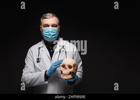 medico senior in maschera medica e guanti in lattice tenendo il cranio isolato su nero Foto Stock