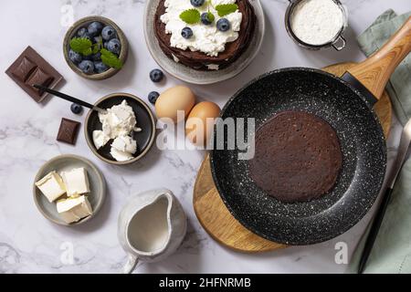 Colazione sana. Ingredienti per la cottura e deliziose crepes con mirtilli e ricotta su un piano in marmo. Vista dall'alto, disposizione piatta. Foto Stock