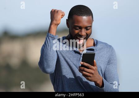 L'uomo eccitato con la pelle nera festeggia le buone notizie controllando il telefono intelligente nella montagna Foto Stock