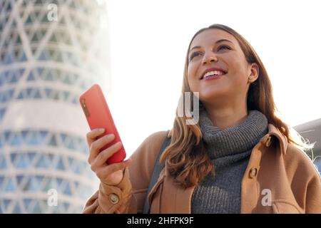 Giovane donna d'affari fiduciosa che guarda al futuro luminoso. Eccitata bella signora che tiene il telefono in mano con moderno grattacielo sul backgro Foto Stock