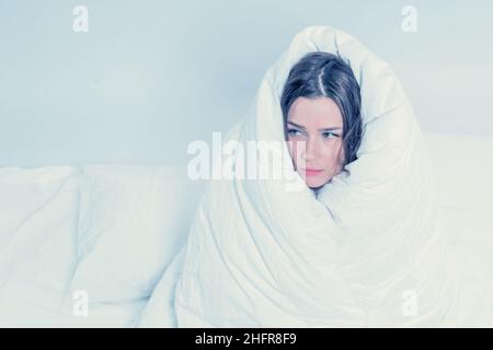 Una graziosa brunetta, avvolta in una coperta, si siede sul letto. Mantenere caldo con una coperta calda. Paura. Eccitamento Foto Stock