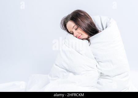 Una graziosa brunetta, avvolta in una coperta bianca di neve, siede sul letto. Mantenere caldo con una coperta calda. Pigrizia. Foto Stock