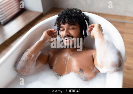 Bell'uomo indiano che indossa cuffie wireless e canta mentre si trova in bagno caldo e frizzante a casa Foto Stock