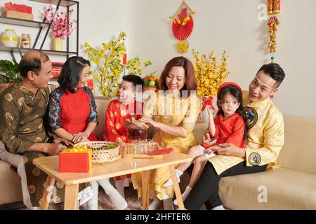 Famiglia di tre generazioni che festeggia il Tet a casa, parlando e gustando il tè con frutta candita Foto Stock