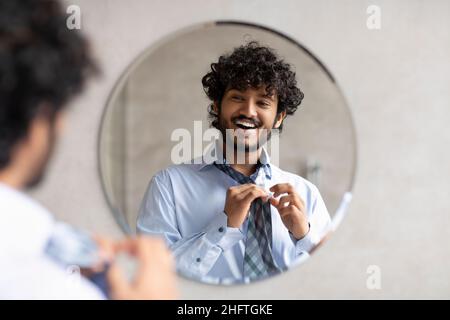 Felice uomo d'affari indiano che mette su cravatta mentre guarda nello specchio, in piedi nella stanza da bagno moderna, spazio libero Foto Stock