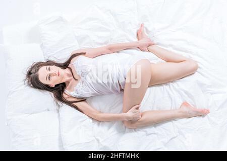 La ragazza si sveglia al mattino facendo yoga a letto, allunga le gambe. Esercizio al mattino. La brunetta conduce uno stile di vita sano. Camera bianca Foto Stock