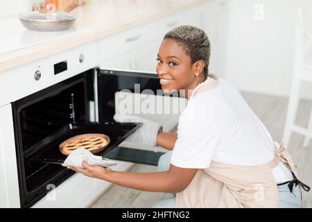 Allegro giovane bella donna nera con capelli corti toglie la torta dal forno in moderno interno minimalista cucina Foto Stock