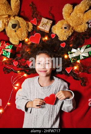 una bambina con una ghirlanda e cuori rossi in capelli ricci sorride su uno sfondo rosso e tiene un cuore nelle sue mani. Foto Stock