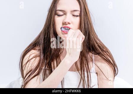 Buongiorno. La ragazza è impegnata in igiene orale. Svegliati presto la mattina. Dorata brunetta spazzolando i denti mentre si siede sul letto con clos Foto Stock