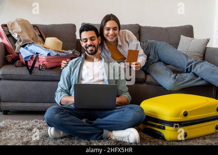 Coppia entusiasta che usa il portatile per prenotare i biglietti e l'hotel online, preparandosi per le vacanze e le valigie d'imballaggio Foto Stock