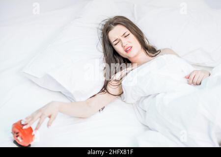 La brunetta si trova, si sveglia dal suono della sveglia. Rumore forte. Letto bianco. Buongiorno. La donna si infastidisce, spegne la sveglia Foto Stock