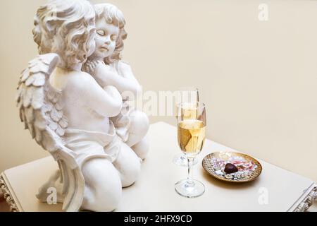 Due bicchieri di champagne e una caramella a forma di cuore su un piatto vicino a due angeli d'amore il giorno di San Valentino. Messa a fuoco soft. Bicchieri di champagne per la sposa Foto Stock