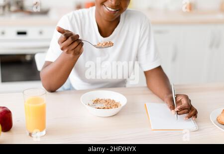 Sorridente femmina nera mangiare porridge a tavola con biscotti, succo e mela, prendere appunti Foto Stock