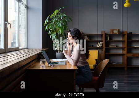 La giovane donna d'affari lavora in remoto su un laptop nello spazio di lavoro, parla con il cliente tramite telefono cellulare Foto Stock